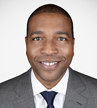 A man smiles at the camera, he has on a suit jacket, collared shirt, and blue tie