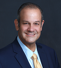 A man smiles at the camera, he has on a suit jacket, collared shirt, and yellow tie