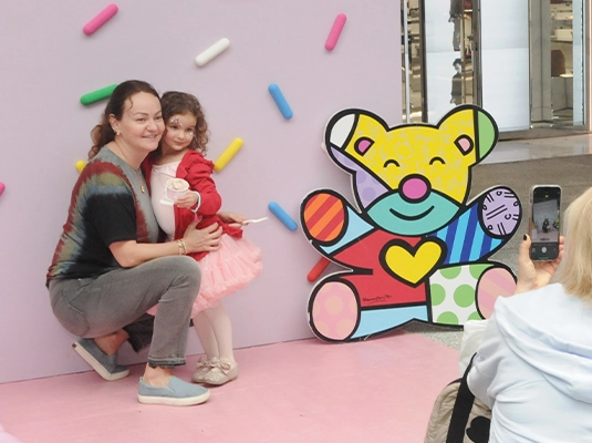 A little girl and a mom posing next to a pink wall and a colorful teddy bear