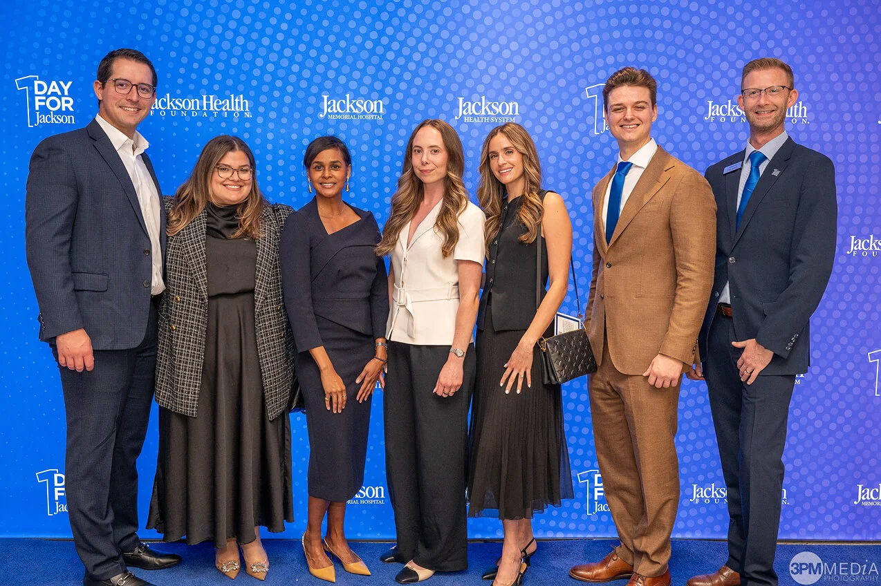 A group of seven professionally dressed young adults posing together in front of a Jackson Health Foundation step-and-repeat banner at a formal event.
