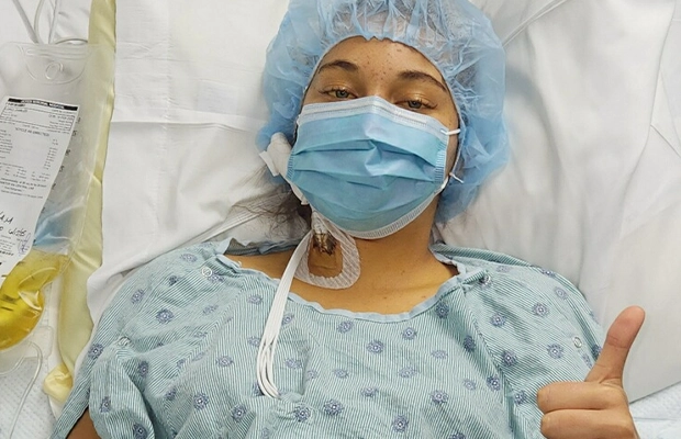 A woman giving a thumbs-up while lying on a hospital gurney, wearing a cap and a surgical mask.
