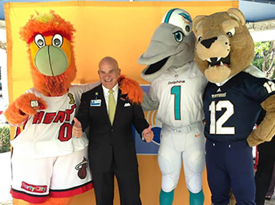 A man smiling as he stands next to three mascots, one mascot is a dolphin, one is a fireball, and a panther