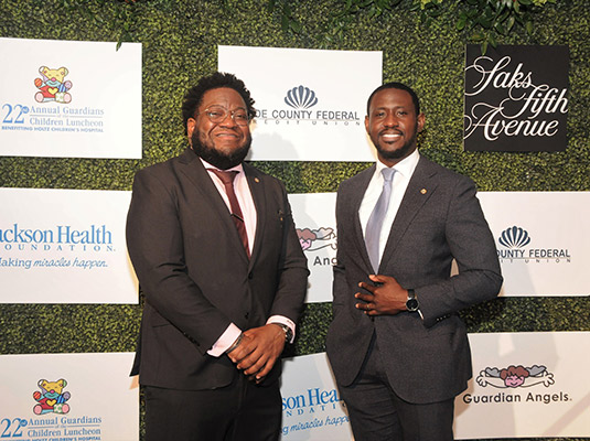 Two men standing in front of a step and repeat, they're smiling at the camera and they both have on suits