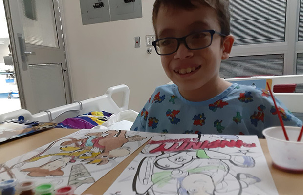 A young boy smiling at the camera, he has coloring pages in front of him