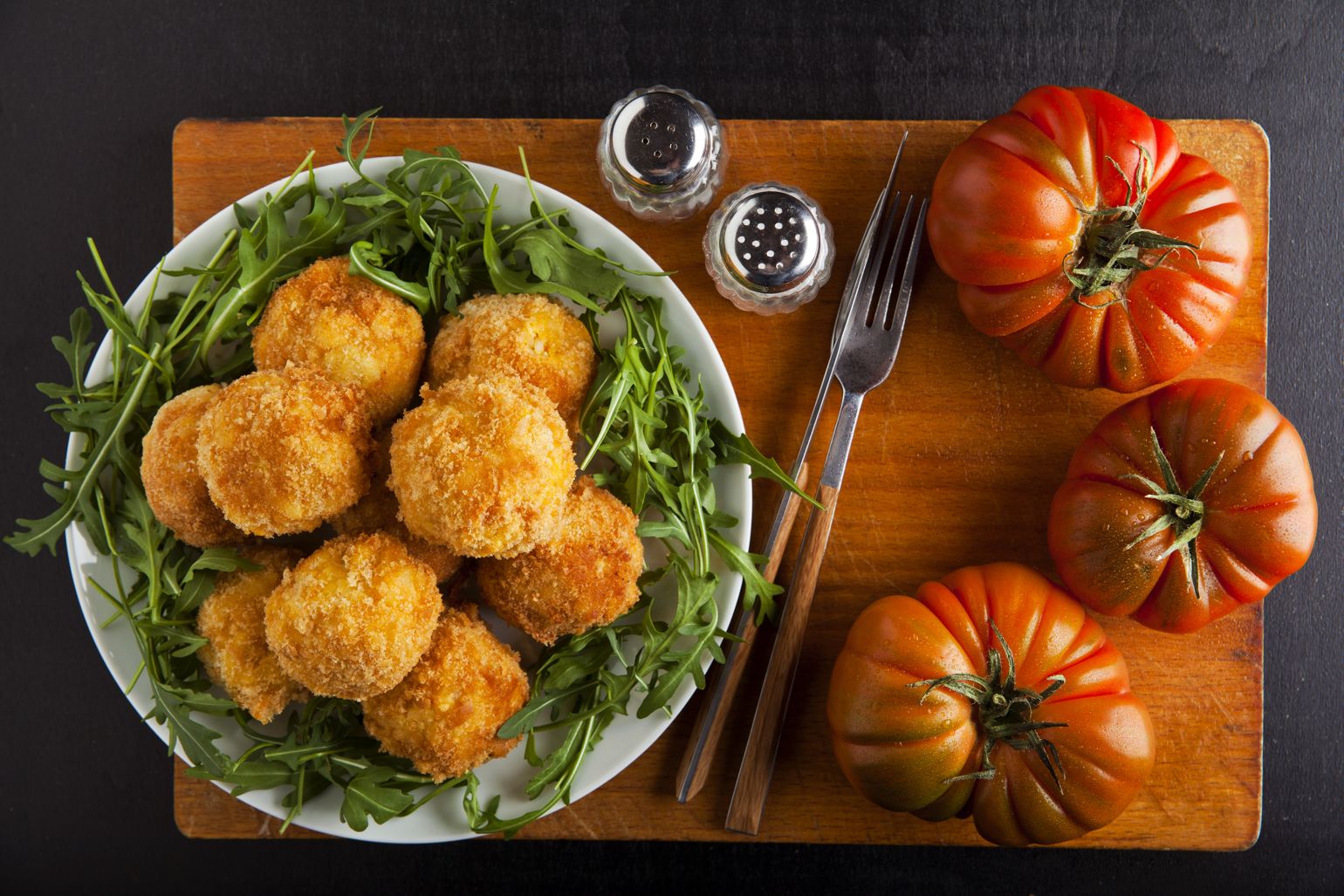 Prato com croquetes de arroz de tomate acompanhados com rúcula, talheres, saleiro, pimenteiro e tomates inteiros sobre tábua de corte de madeira.