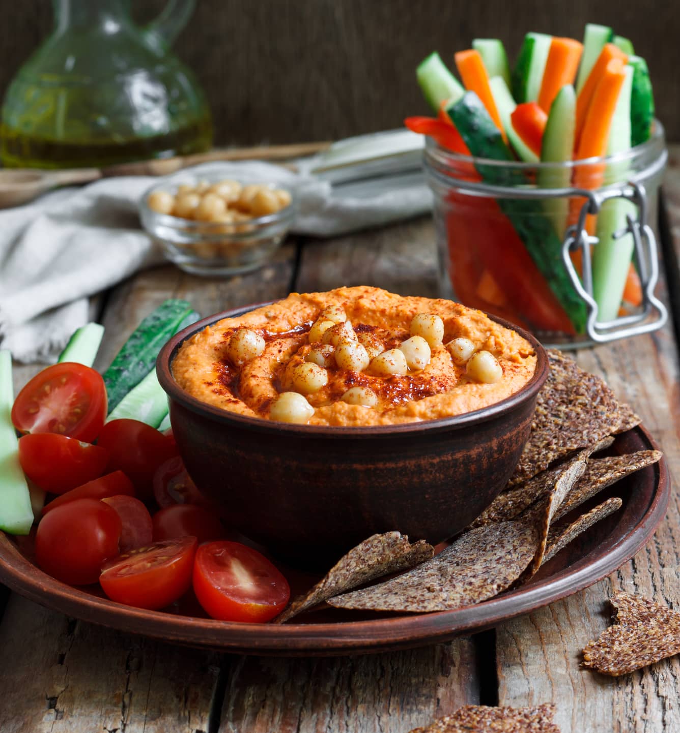 Taça com hummus, acompanhada de palitos de pepino e cenoura, tomates cherry e tostas integrais.