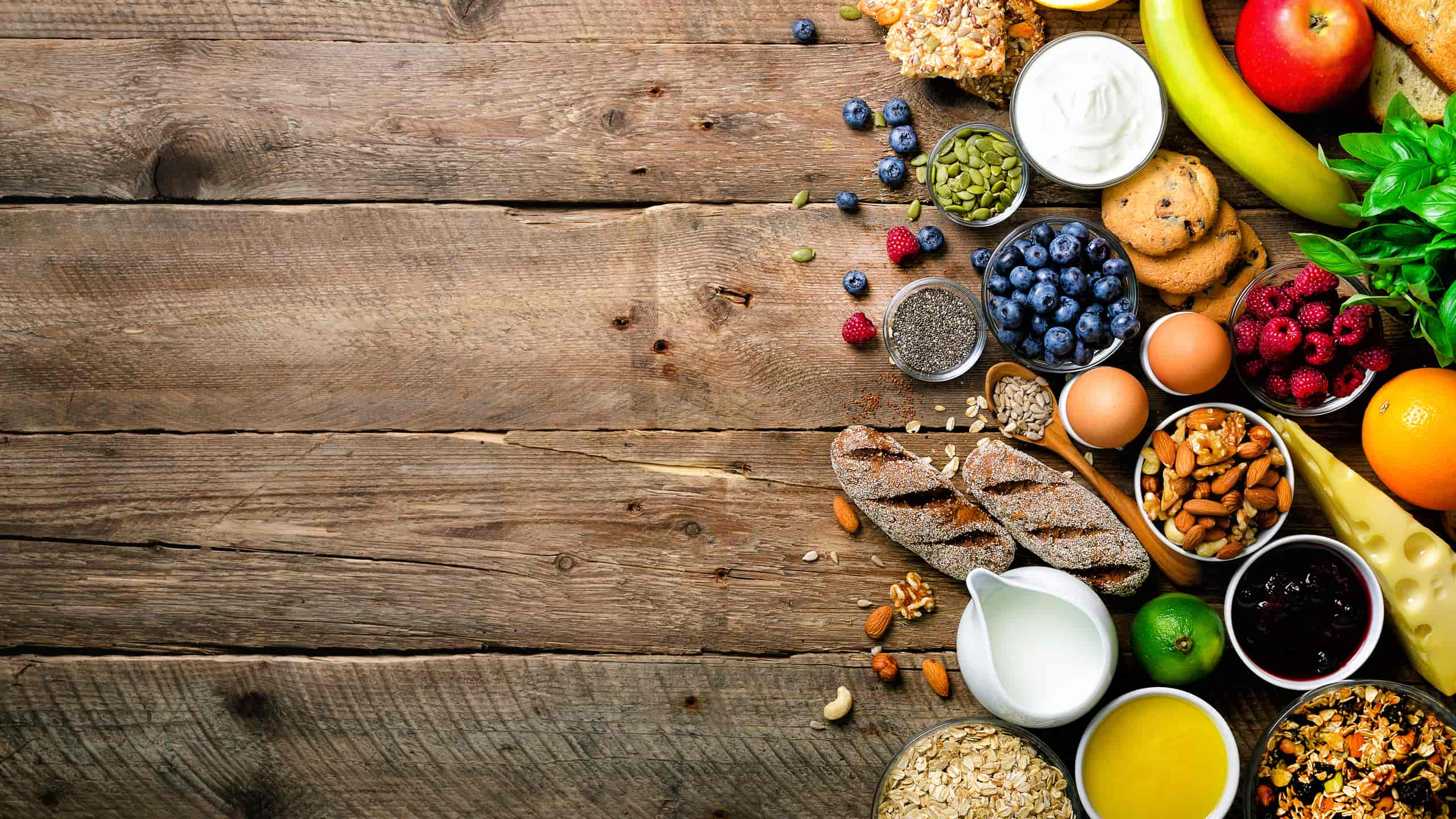 Snacks saudáveis em mesa de madeira: barras de cereais, iogurte, banana, maçã, laranja, lima, pevides, bolachas, mirtilos, sementes de chia, framboesas, ovos, frutos secos, pão integral, leite, queijo, granola e aveia.