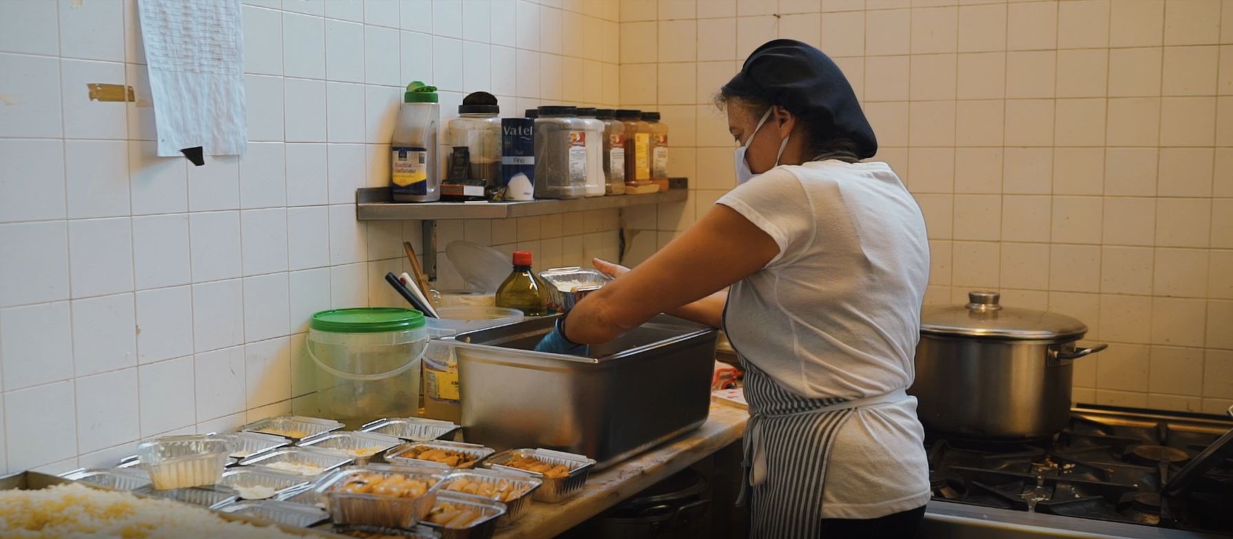 Mulher em cozinha, junto a fogão, colocando refeições em recipientes individuais de alumínio.