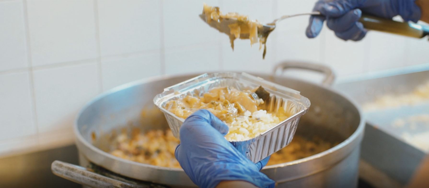 Mão retirando comida de uma panela com uma colher e colocando em recipiente individual de alumínio.