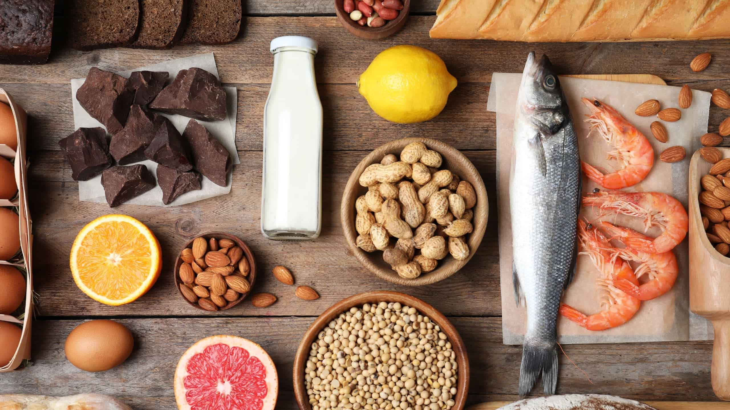 Table with several foods: chocolate, milk, lemon, peanuts, almonds, fish, seafood, eggs, orange, bread.