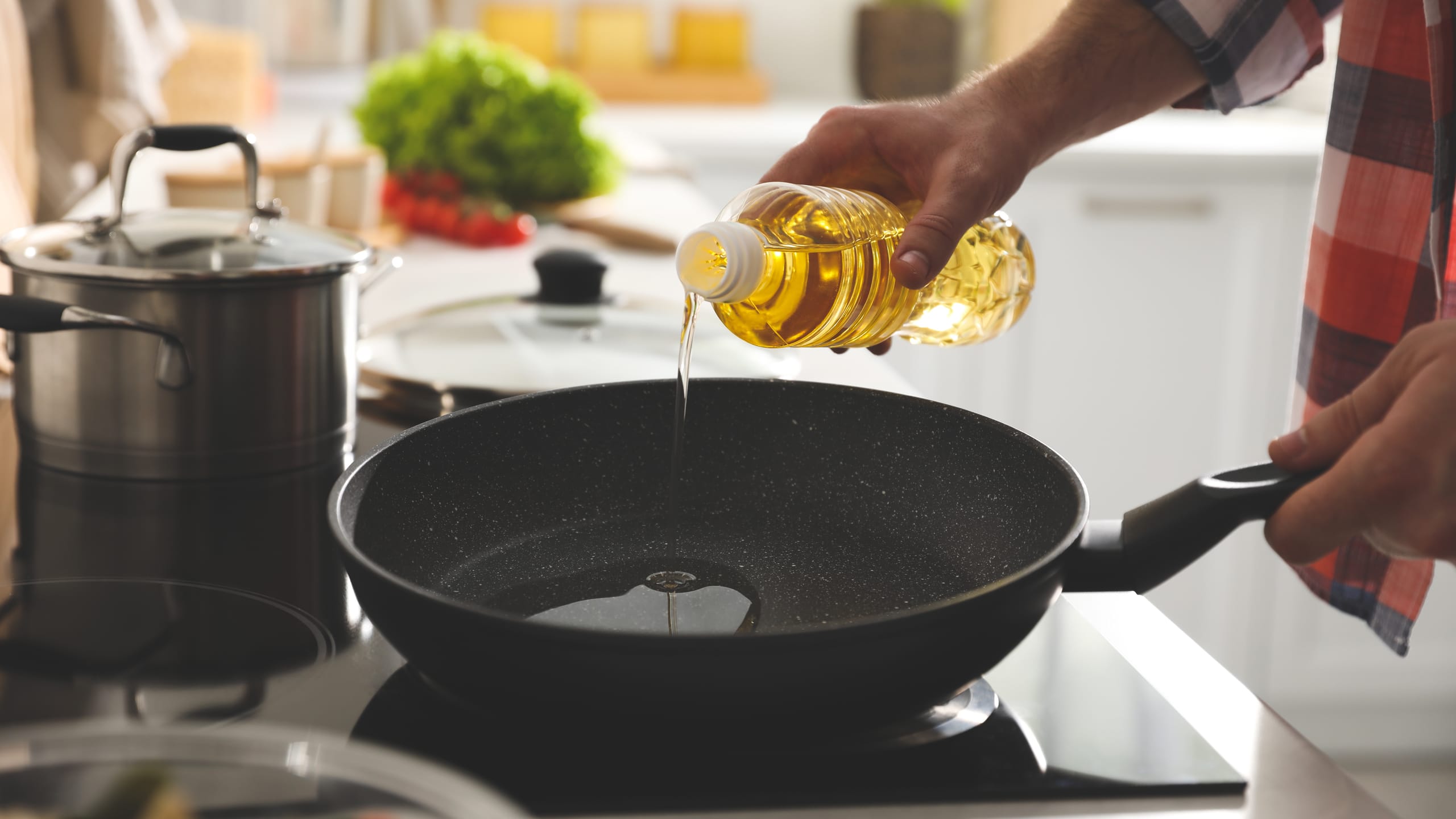 Mão masculina despejando óleo alimentar numa frigideira, em placa elétrica na cozinha.