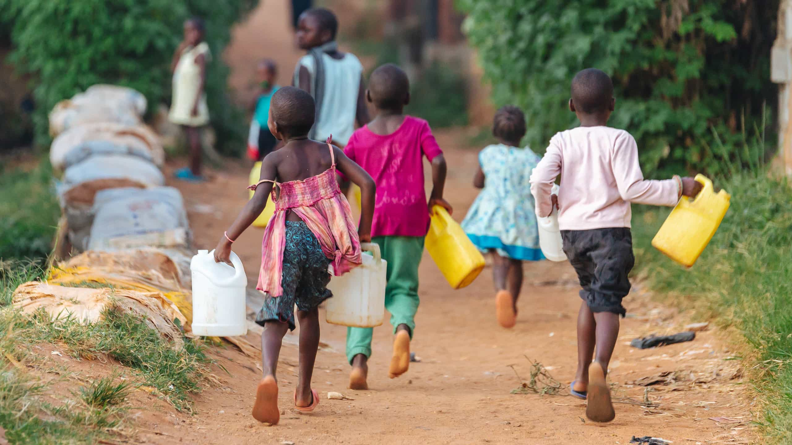 Crianças a correr em caminho de terra batida, segurando garrafões para recolher água.