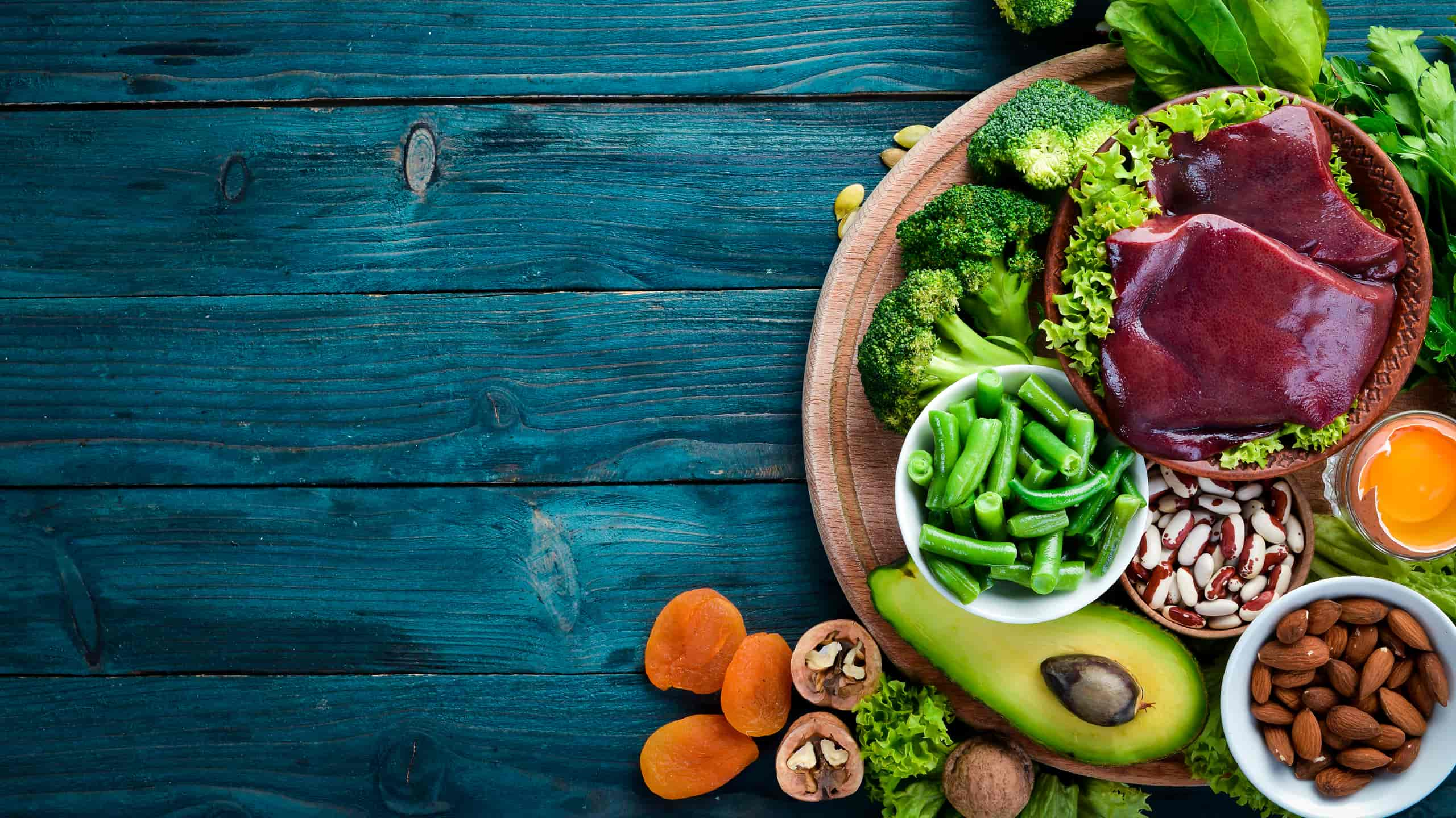 Tábua de madeira com alimentos com ferro: brócolos, espinafre, feijão-verde, fígado, feijão, amêndoas, abacate, ovos, nozes, alperces.