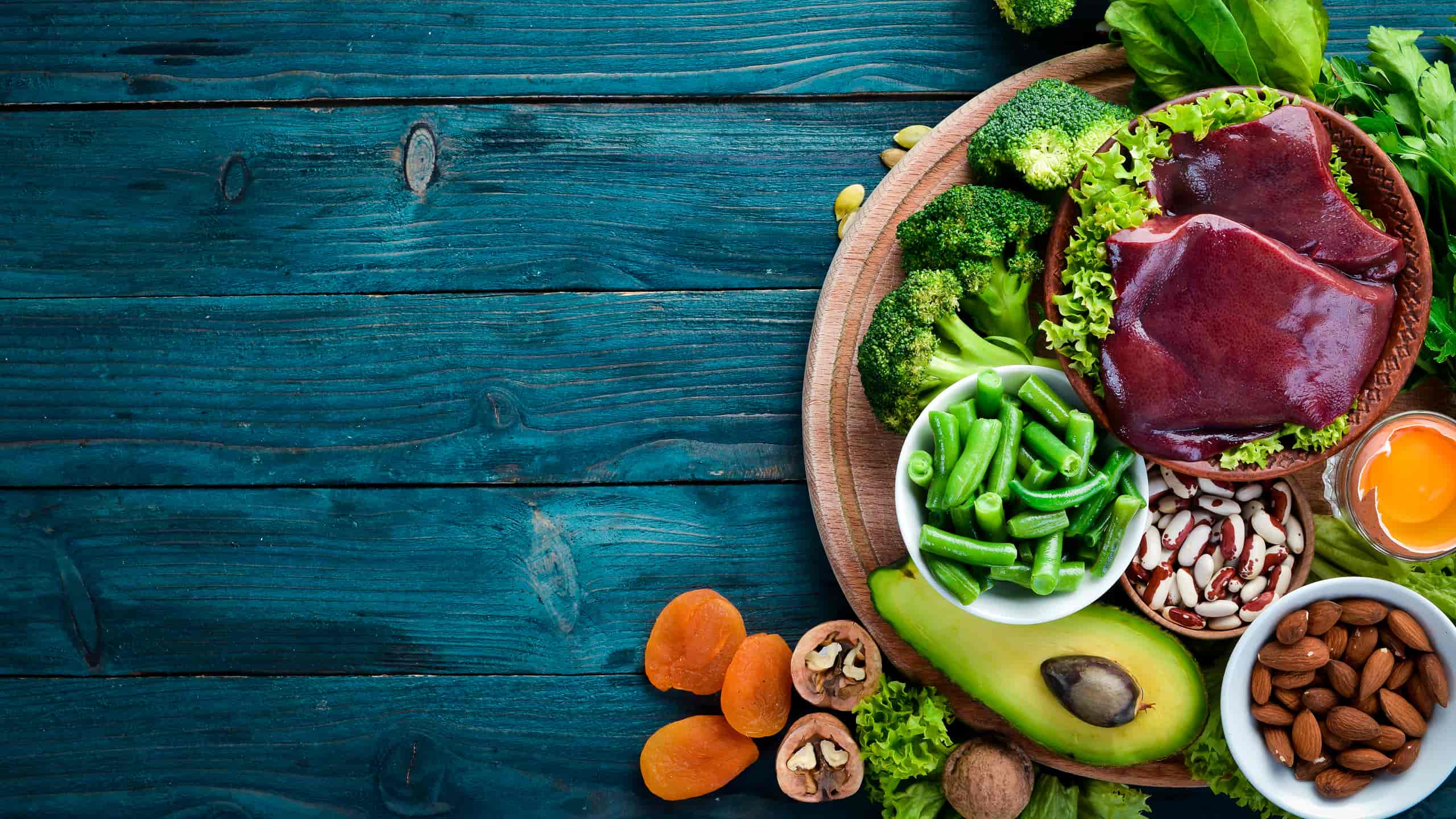 Wooden board with high in iron foods: broccoli, green beans, liver, beans, almonds, avocado, egg, walnuts and apricots.