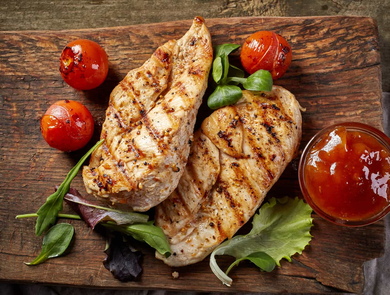 Dois peitos de frango grelhados, tomate cherry e folhas de alface, em tábua de corte de madeira.