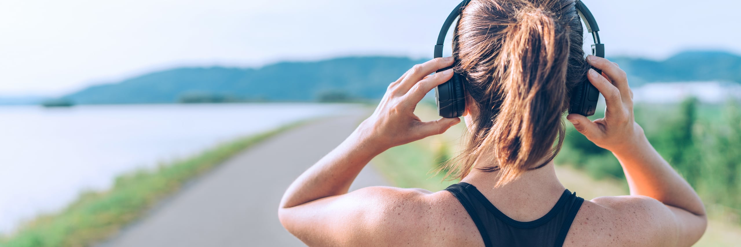 Mulher ajustando headphones, com roupa desportiva, em estrada à beira rio.