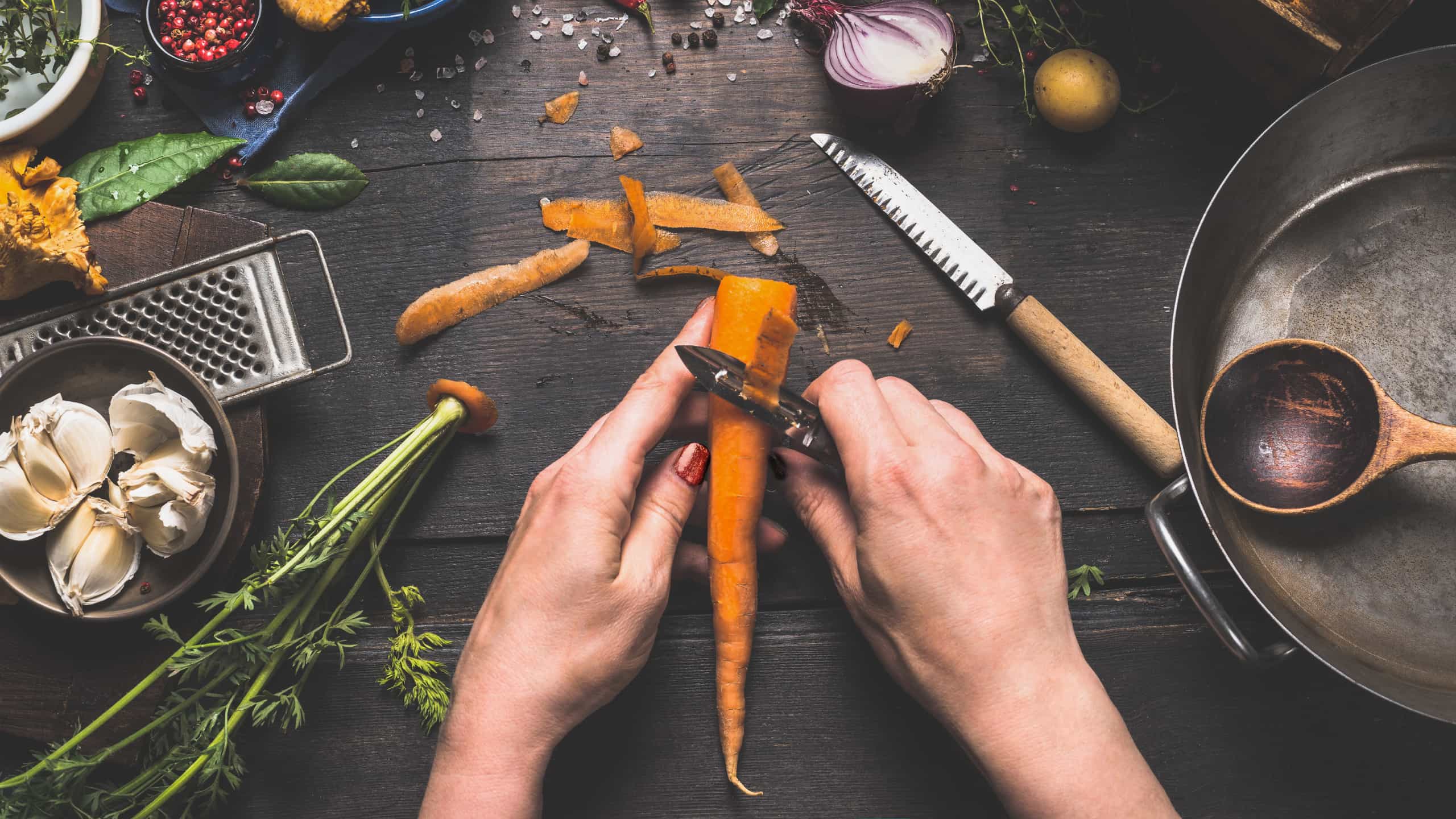 Mãos femininas a tirar a pele da cenoura utilizando um descascador, em mesa de madeira com legumes e vários utensílios de cozinha.