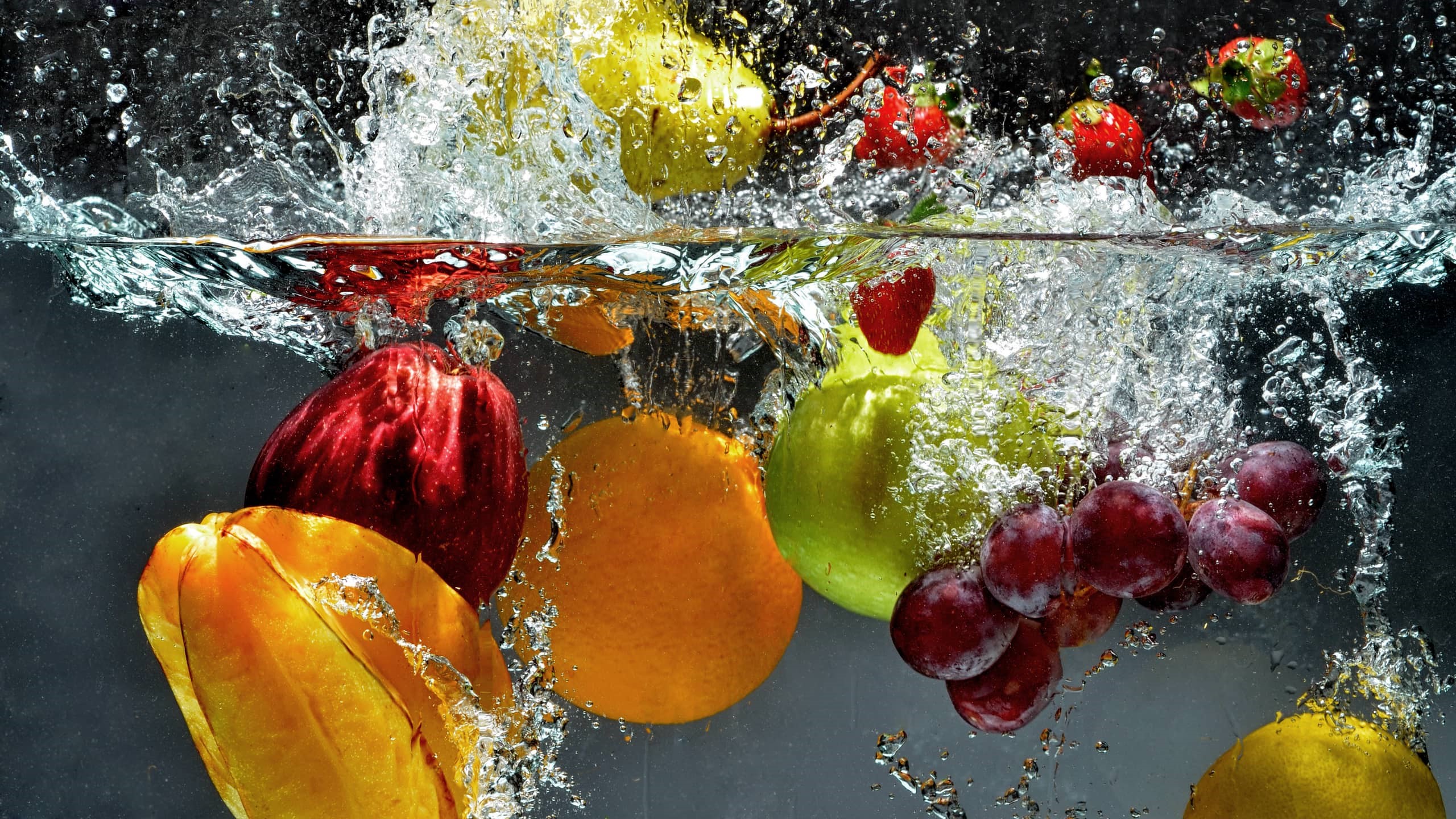 Several fruits falling in the water: apple, orange, pear, strawberries, grapes.