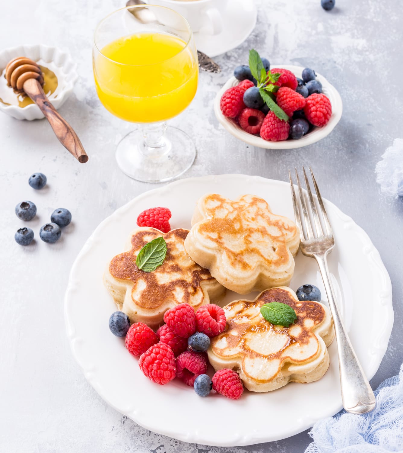 Plate with flower-shaped pancakes served with blueberries and raspberries and a glass of fruit juice.