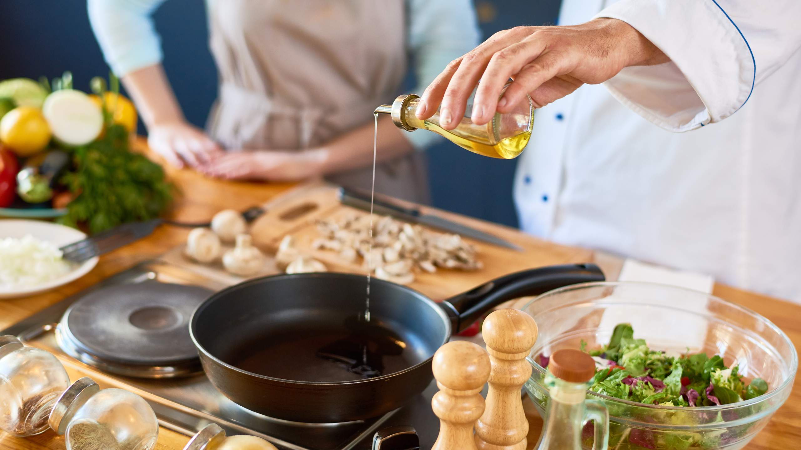 Mão de homem com jaleca, vertendo óleo sobre uma frigideira que se encontra numa placa portátil elétrica sobre bancada de cozinha com vários alimentos e utensílios.