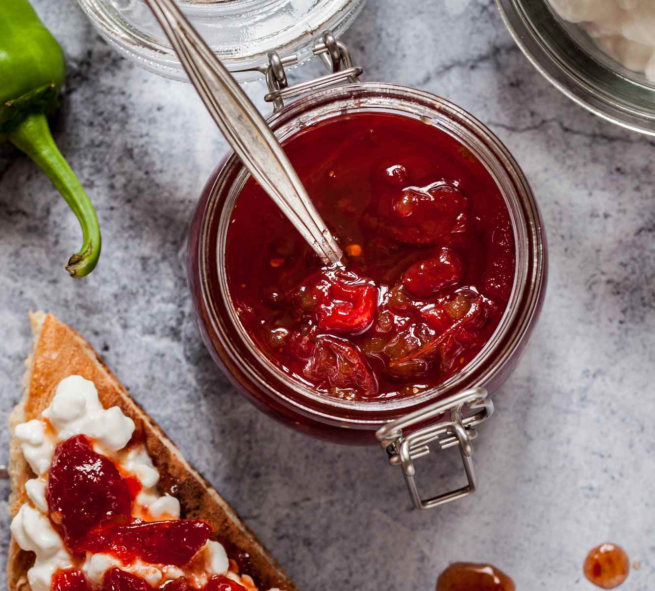 Frasco de chutney caseiro de tomate com colher, e tosta com ricotta e chutney, sobre tampo de mármore.