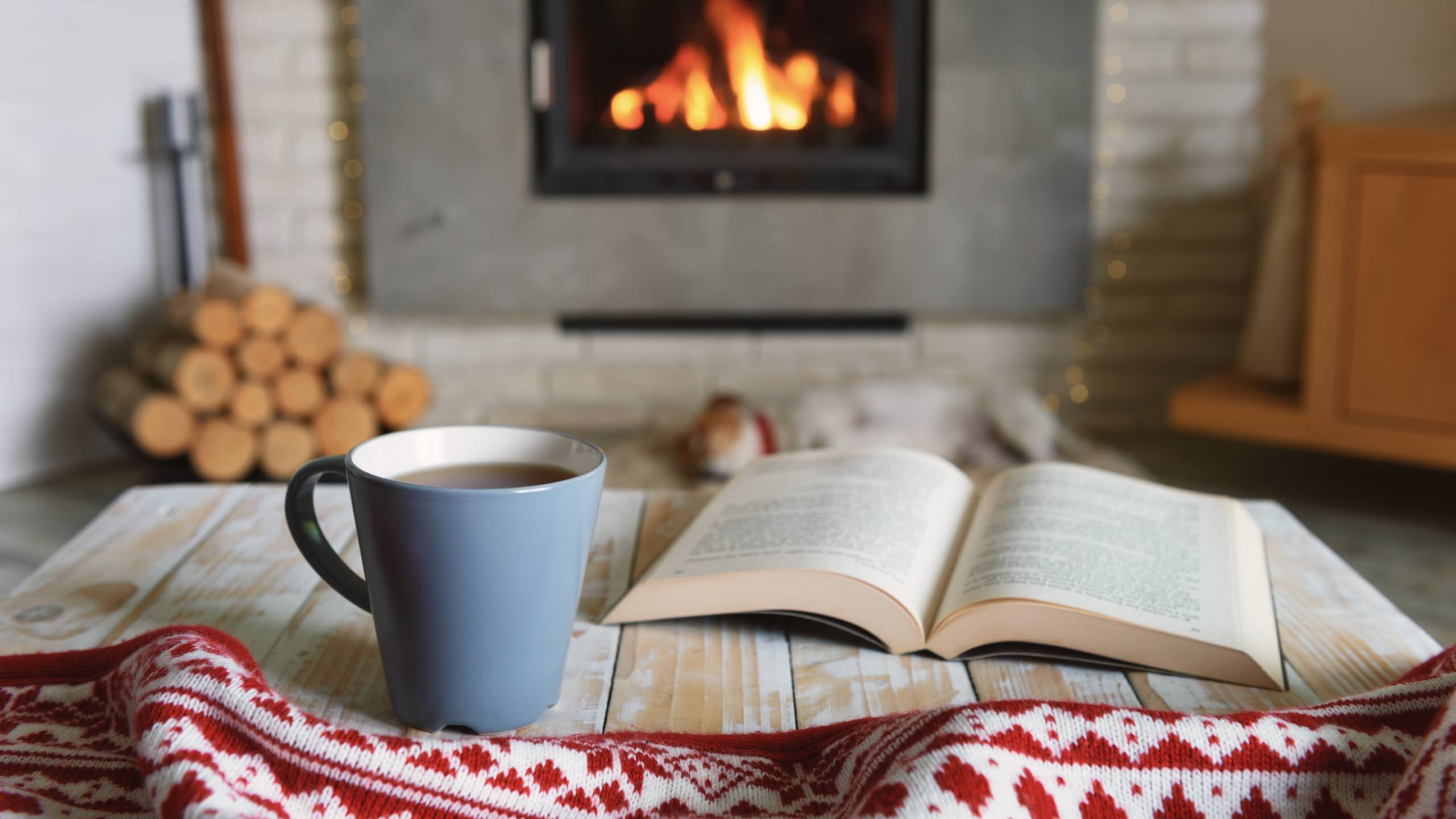 Blanket, cup of tea and open book on a wooden table, in a room with a lit fireplace.