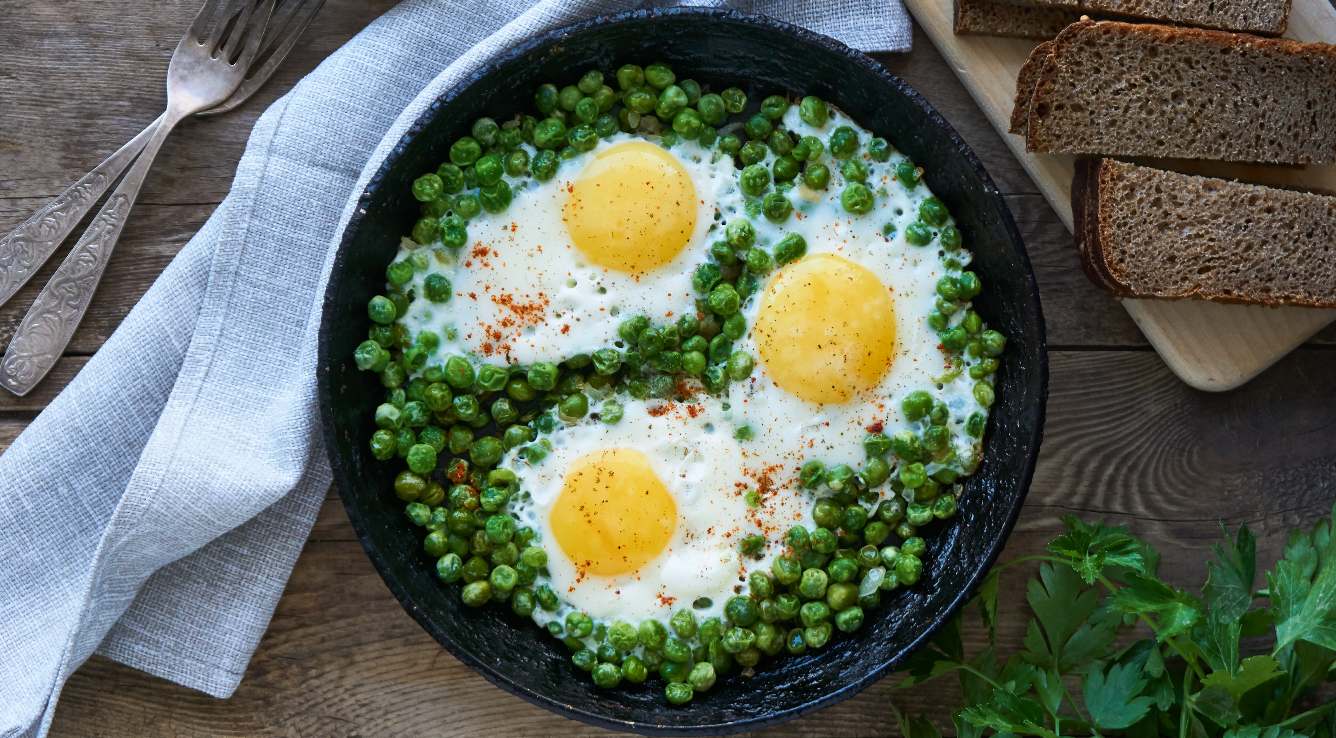 Deep plate with green peas with poached eggs, whole bread slices, cloth and forks over a wooden table.