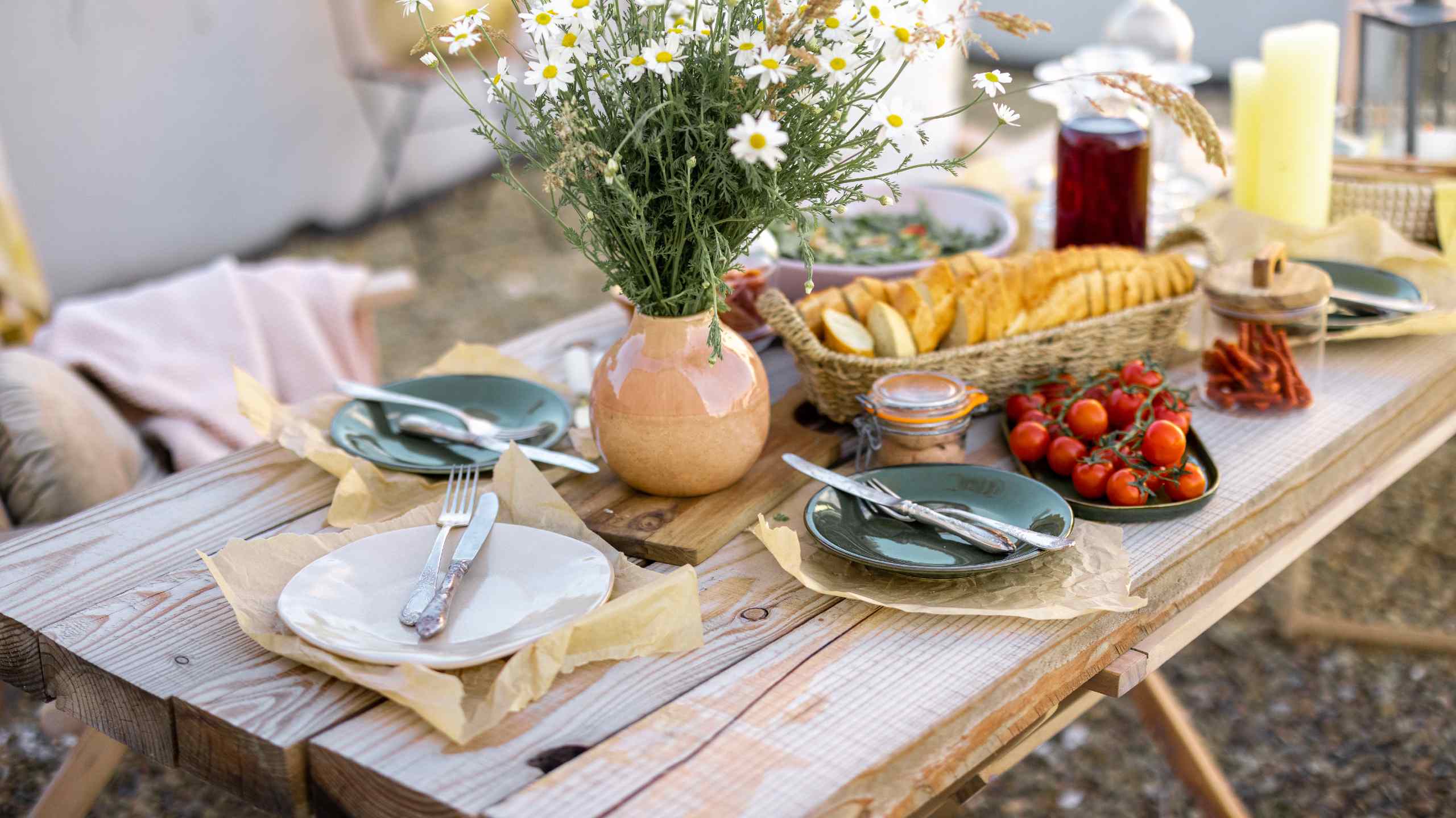 Mesa exterior posta para almoço, com cinco pratos e talheres, cesto de pão, prato com tomates cherry e jarra cerâmica com malmequeres.