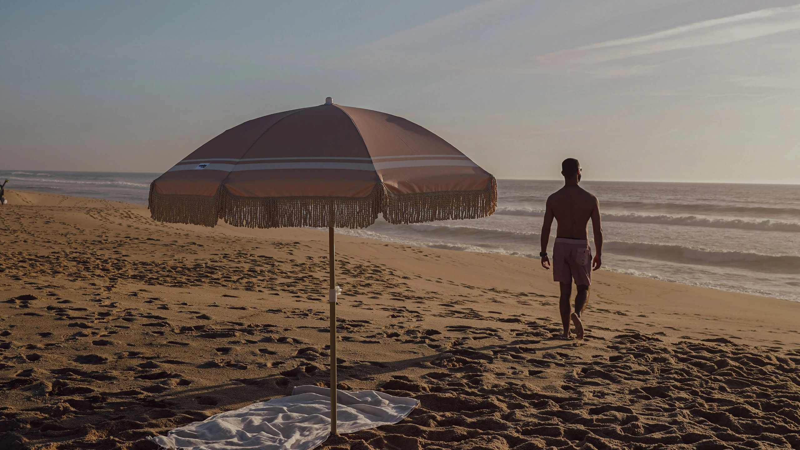 Chapéu de sol Sombra & Co colocado na areia na praia, junto a toalha.