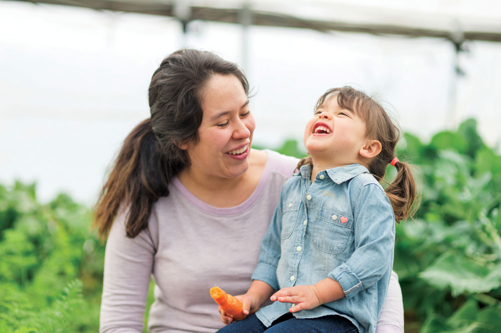 Young ethnic mom holds her daughter on lap who is snacking on a carrot from the garden. She is leaning her head back and laughing uproariously.