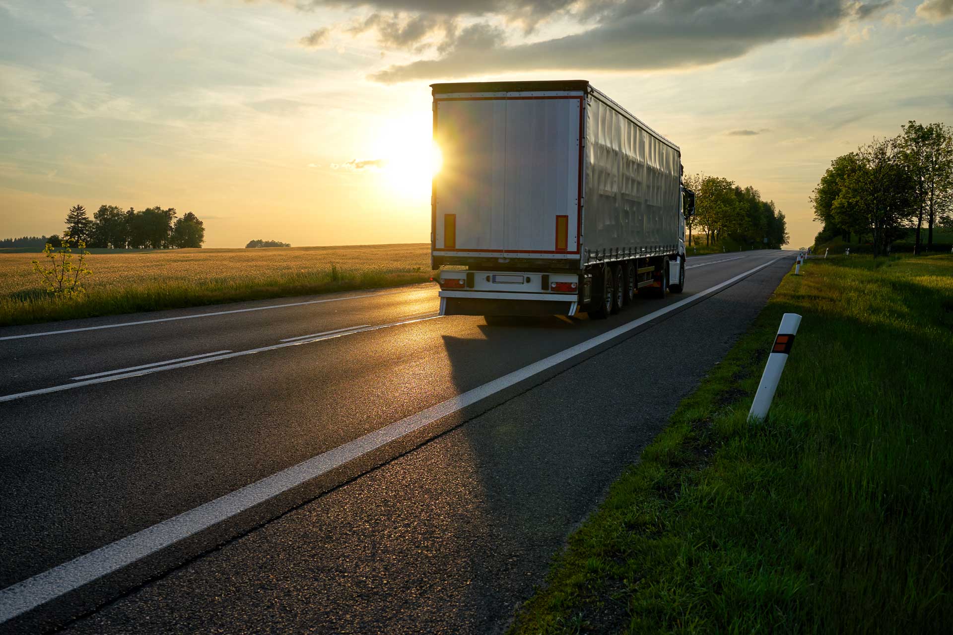 "Truck departing towards the horizon on an asphalt road in a rural countryside at sunset. "