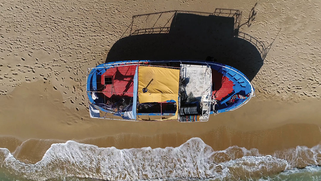 Aerial top down photo of beach and stranded refugee boat at southern mediterranean beach showing the colorful vessel located at the yellow beach with human footprints also showing the azure blue ocean