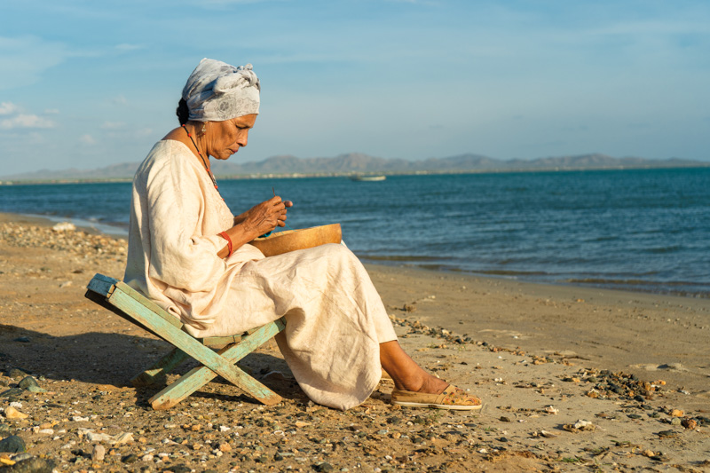 elderly woman sitting on a chair by the sea, on a sunny day, sewing a bag