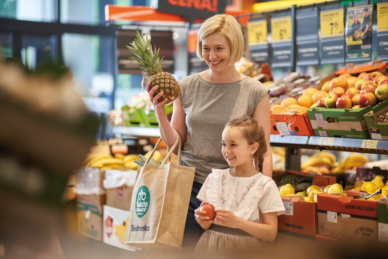 Image of a young woman in the fruits corridor of a Biedronka's store, holding a pineapple, with a young kid by her side holding an apple