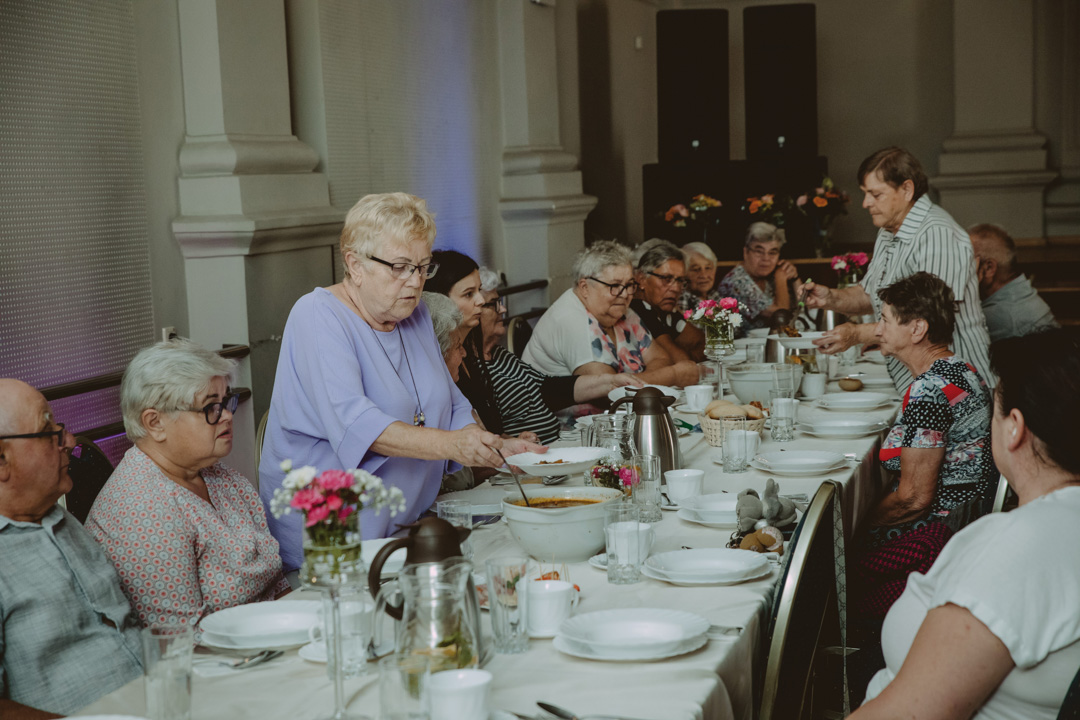 image of a long table filled with elderly women sitted with plates in front of them, and of of them is pouring soup into a plate
