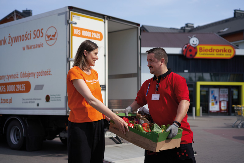 Image of a male biedronka's employee giving a female association worker a box filled with vegetables, outside a biedronka's store