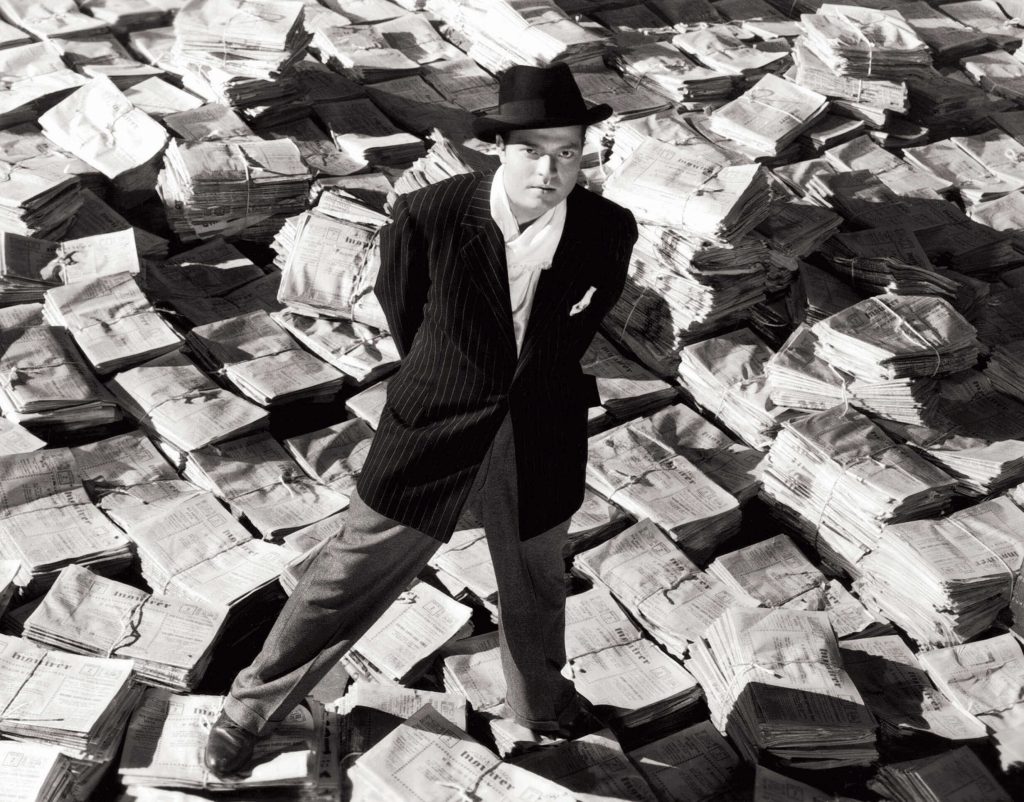  black and white frame from the movie Citizen Kane, by Orson Welles, where a man with a hat dressed in a suit is standing among a lot of newspapers, looking up.