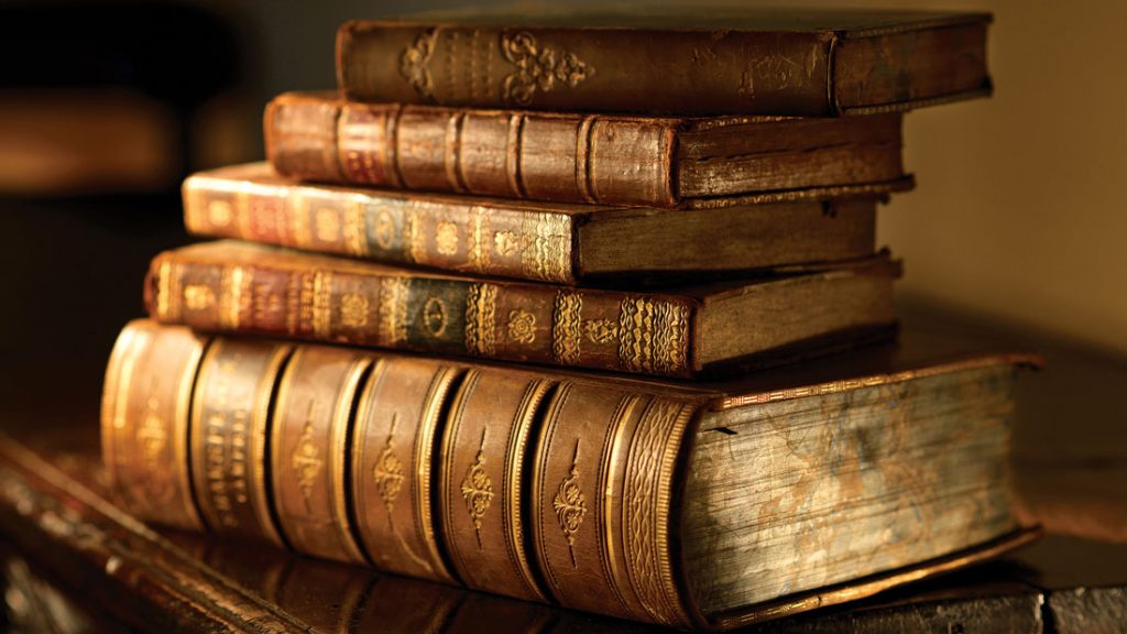 Vintage, antiquarian books pile on wooden surface in warm directional light. Selective focus.