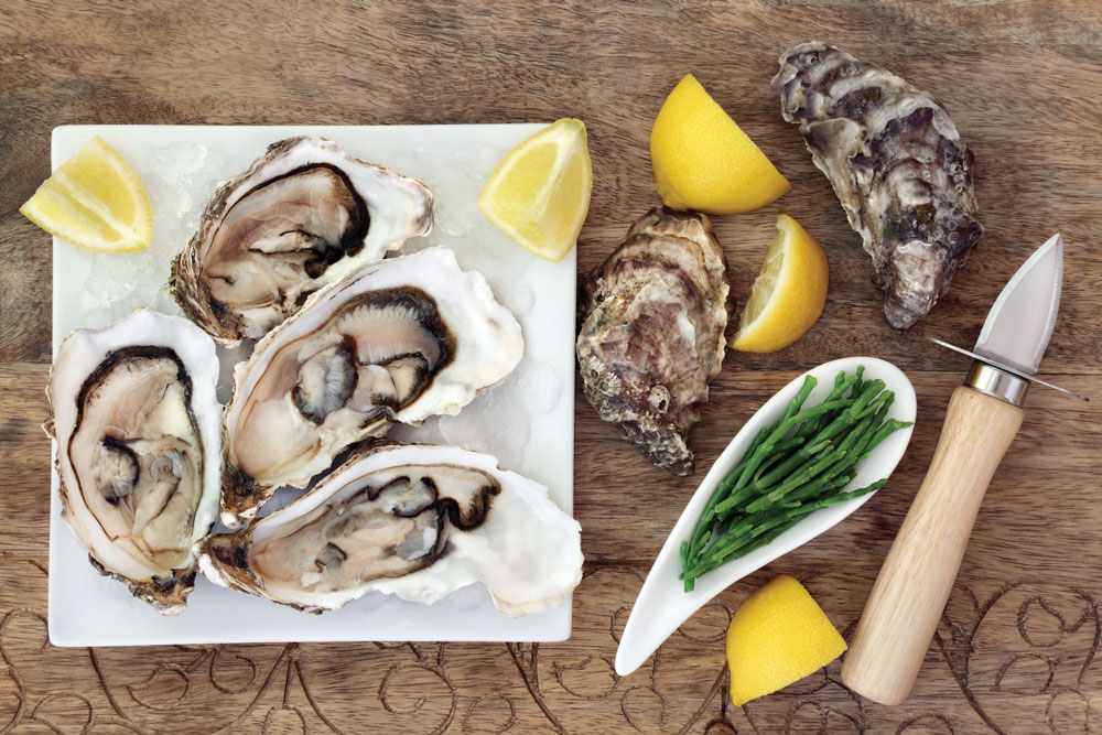 Oysters on crushed ice on a square china plate with oyster knife, samphire and lemon fruit on old wooden background