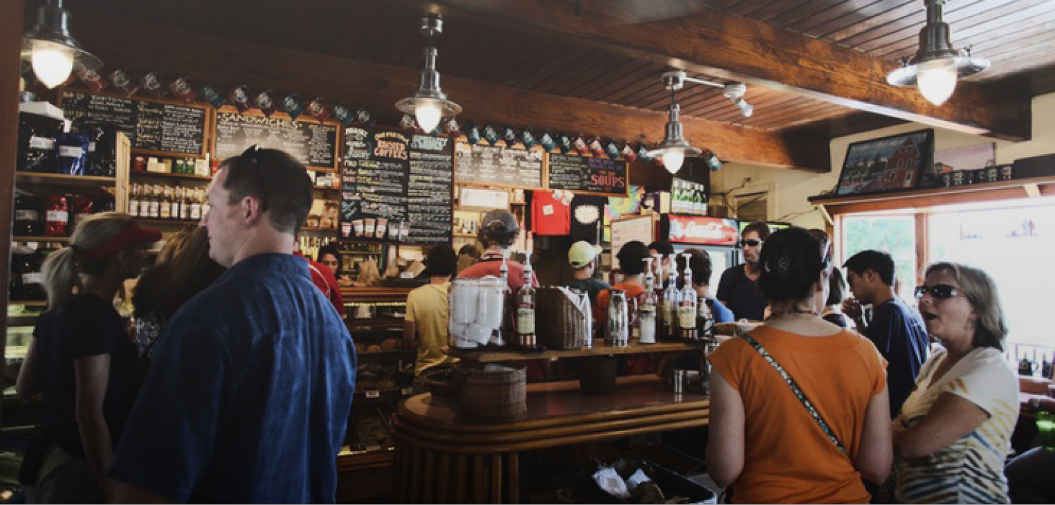 Photograph showing a busy cafe-bar. There are many hand written menus on the back wall, and lots of people queuing and chatting.