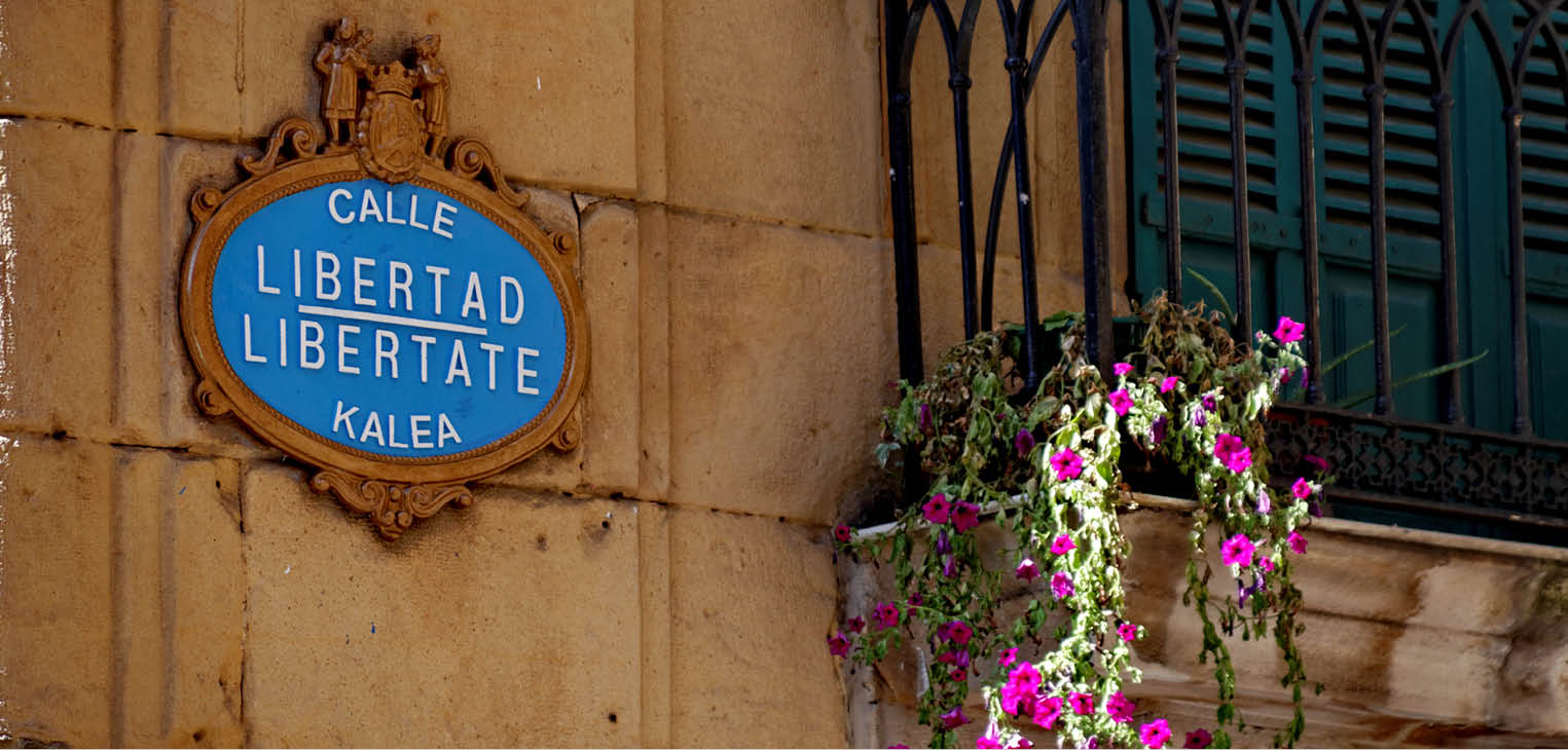 Photograph of an ornate, oval blue bilingual street sign in Spanish and Basque on a yellow stone wall next to a wrought iron balcony with flowers.
