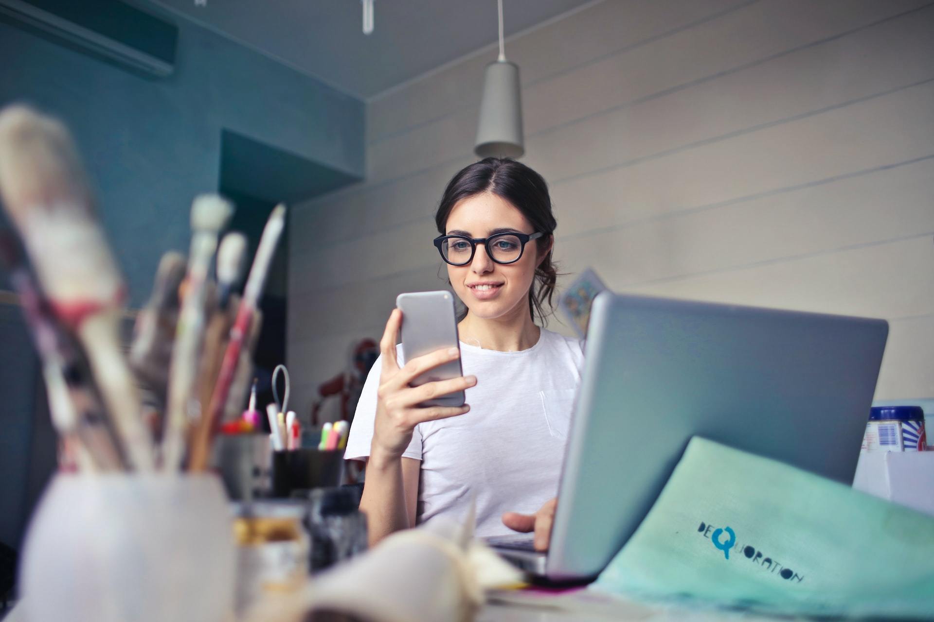 A woman wearing glasses looking at her phone next to a table with art supplies.