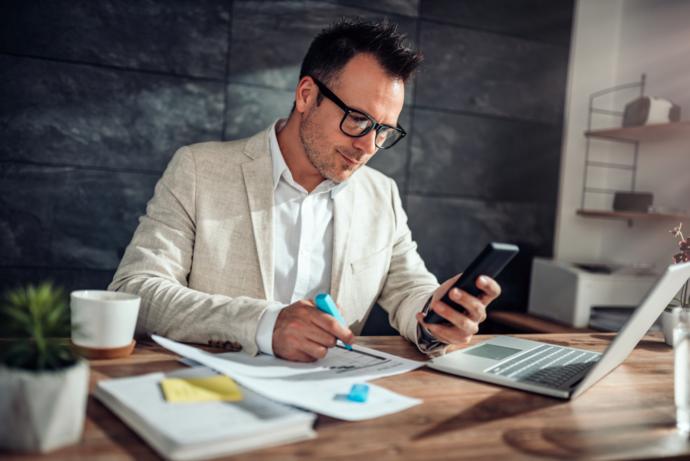 Businessman wearing linen suit and eyeglasses sitting at his desk using smart phone and highlighting text with a blue marker in his office.