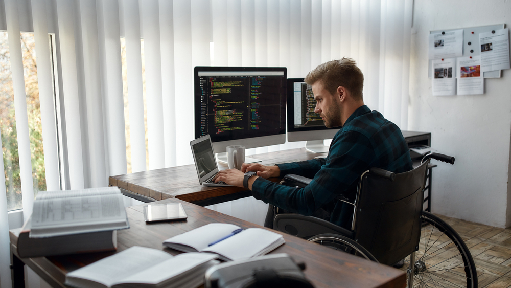 Young caucasian male web developer in a wheelchair writing program code while sitting at workplace with three monitors in the modern office.