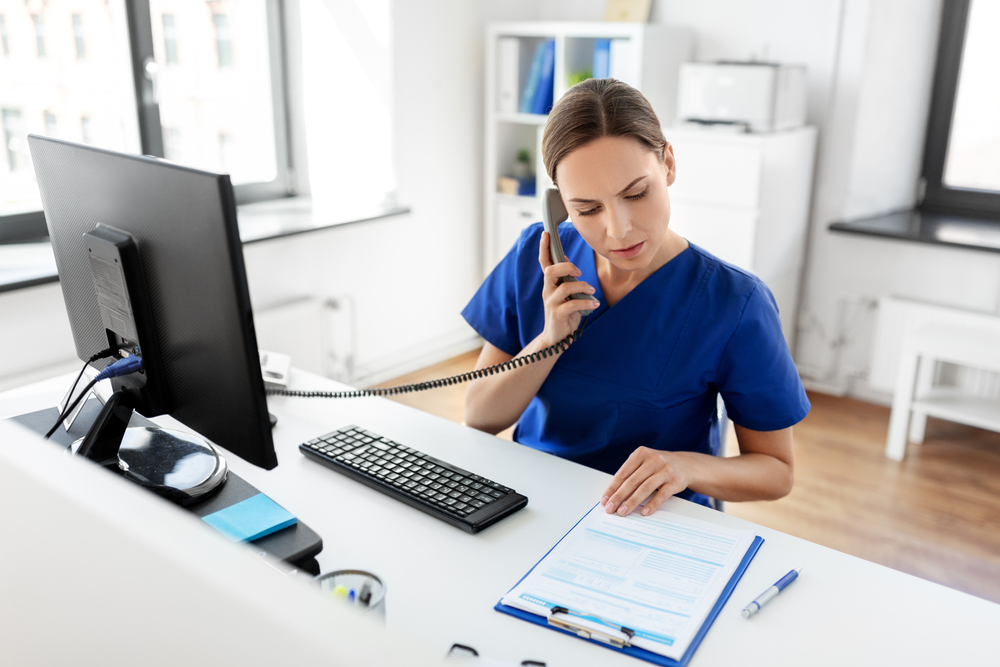 Female doctor or nurse with computer and clipboard calling on phone at hospital.