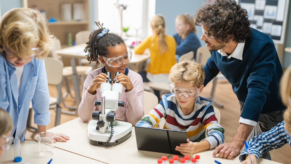 Elementary school science classroom: little girl looks under microscope, boy uses digital tablet to check information on the internet, teacher observes from behind.