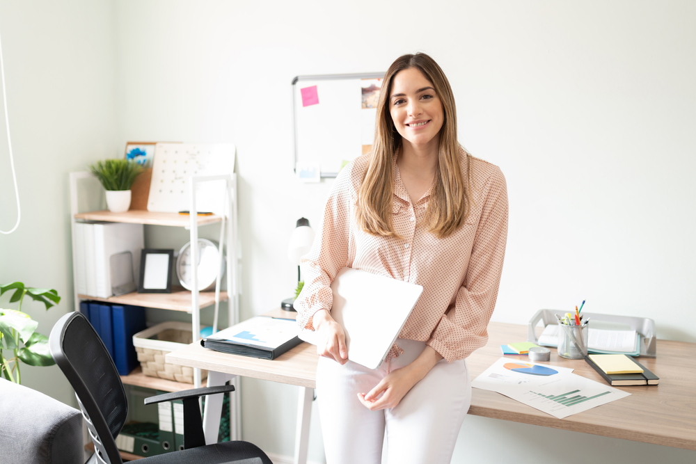 Young woman working as an administrative assistant holding a laptop computer in an office and looking ready to work.