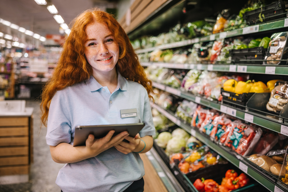 Closeup of a young worker with digital tablet in grocery store; happy trainee working in supermarket.