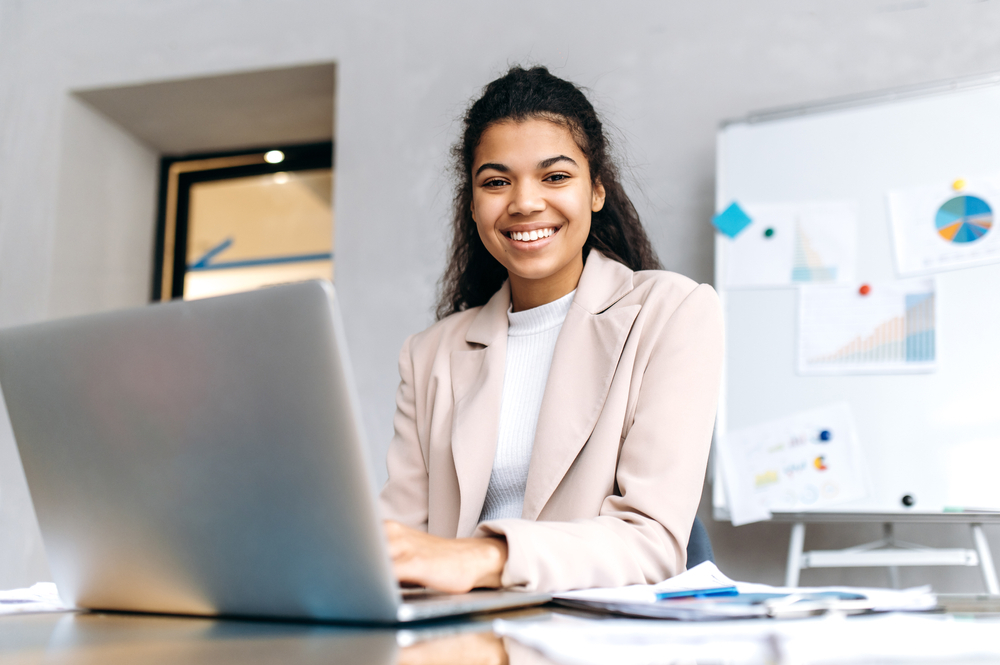 Stylish young business lady sit at the desk, friendly smiling and looks at the camera.