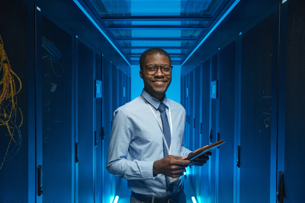 Bespectacled man in a formal office attire including a tie standing in a server cabinet while holding a tablet in an office data center.